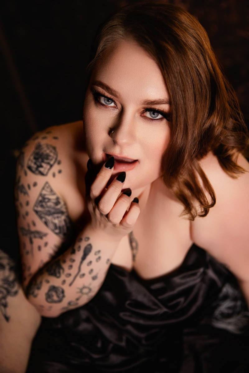 Woman with tattoos posing in black lingerie with finger near lips, intimate boudoir portrait