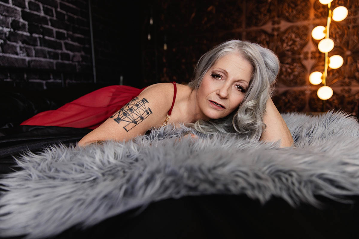 Woman lying on bed in red lace gown with her hand in her silver hair, boudoir photo