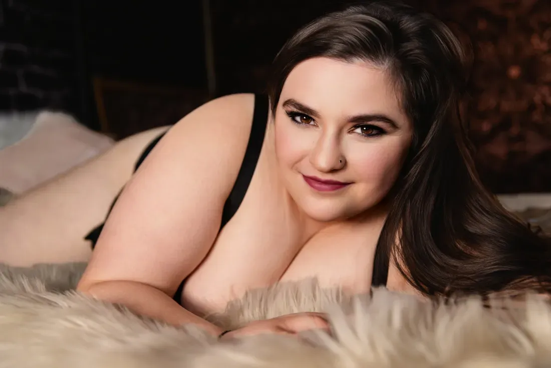 Boudoir portrait of a woman in black lingerie lying on a fur blanket with warm lighting and a confident expression