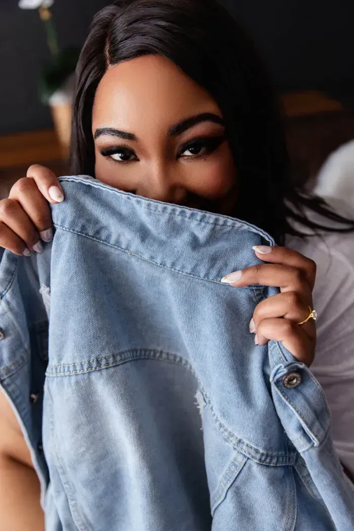 Woman holding denim jacket over face with playful expression during boudoir session
