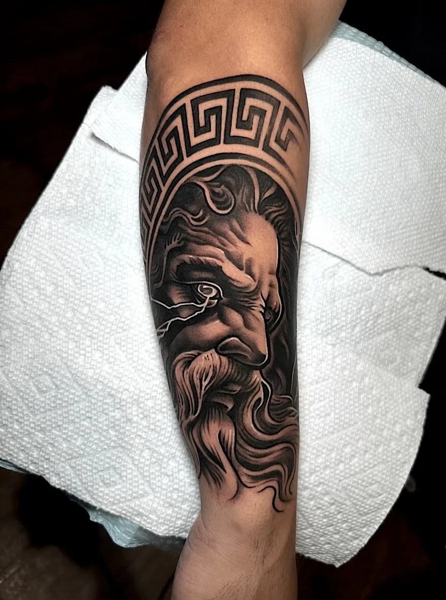 Black and grey bearded classical portrait tattoo beneath a Greek key border