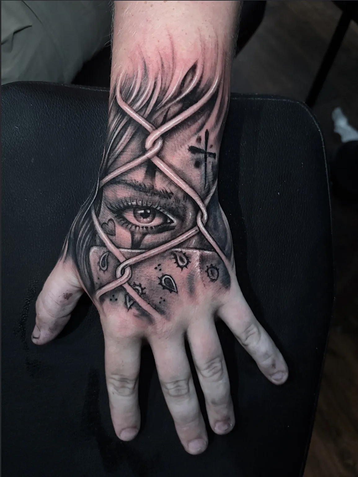 Black and grey hand and wrist tattoo of a woman’s eye behind chain-link fencing by Eric Hooven