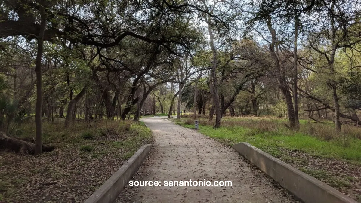 A paved park trail with concrete borders winding through a dense, wooded forest area with tall mature trees, shaded foliage, and scattered leaves on the path. Text at the bottom reads "source: sanantonio.com".