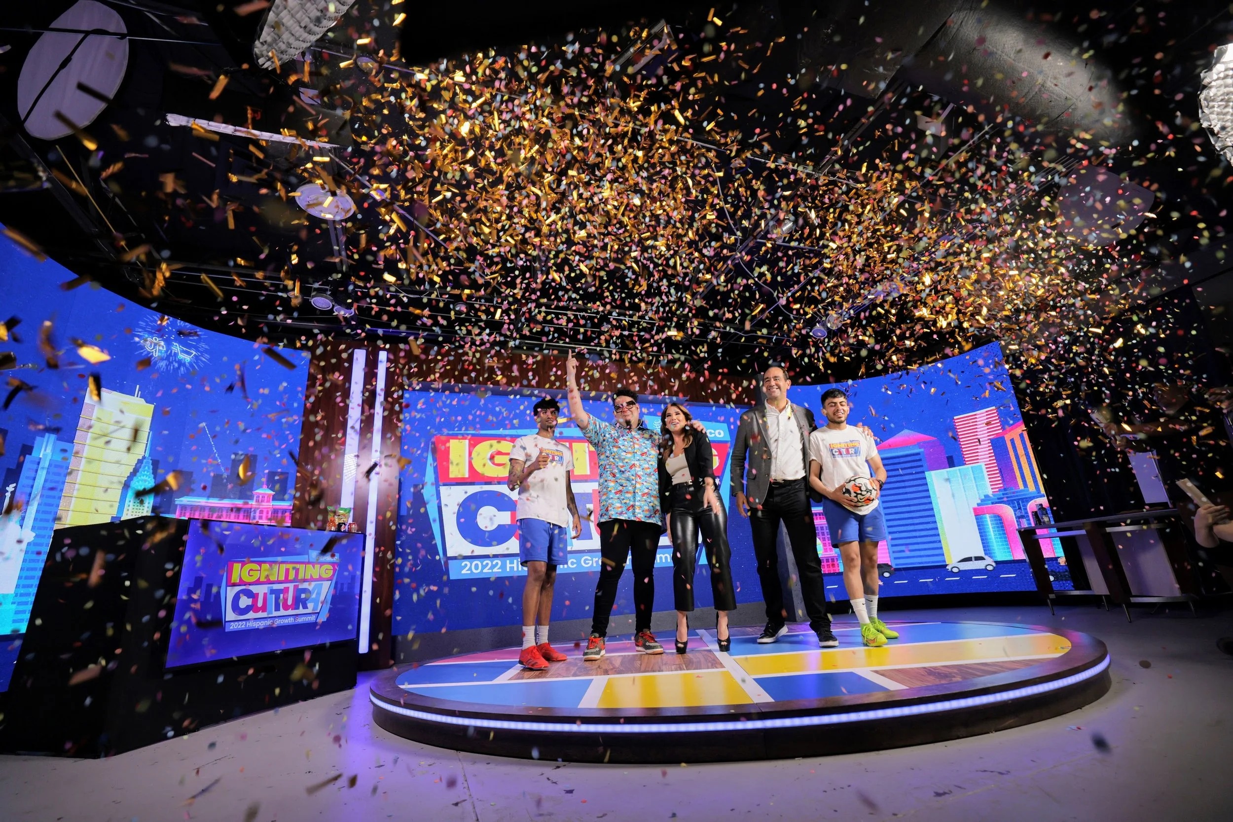 Talk show hosts standing on a stage with an LED screen background while confetti falls.