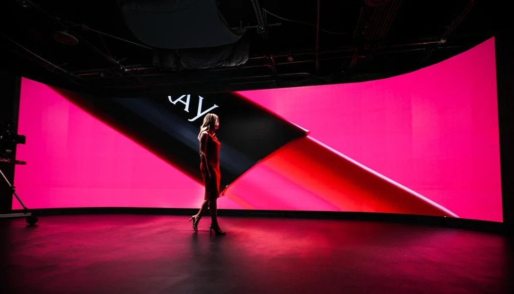Wide shot of a person standing on a stage with a massive glowing pink 'V' graphic behind them.