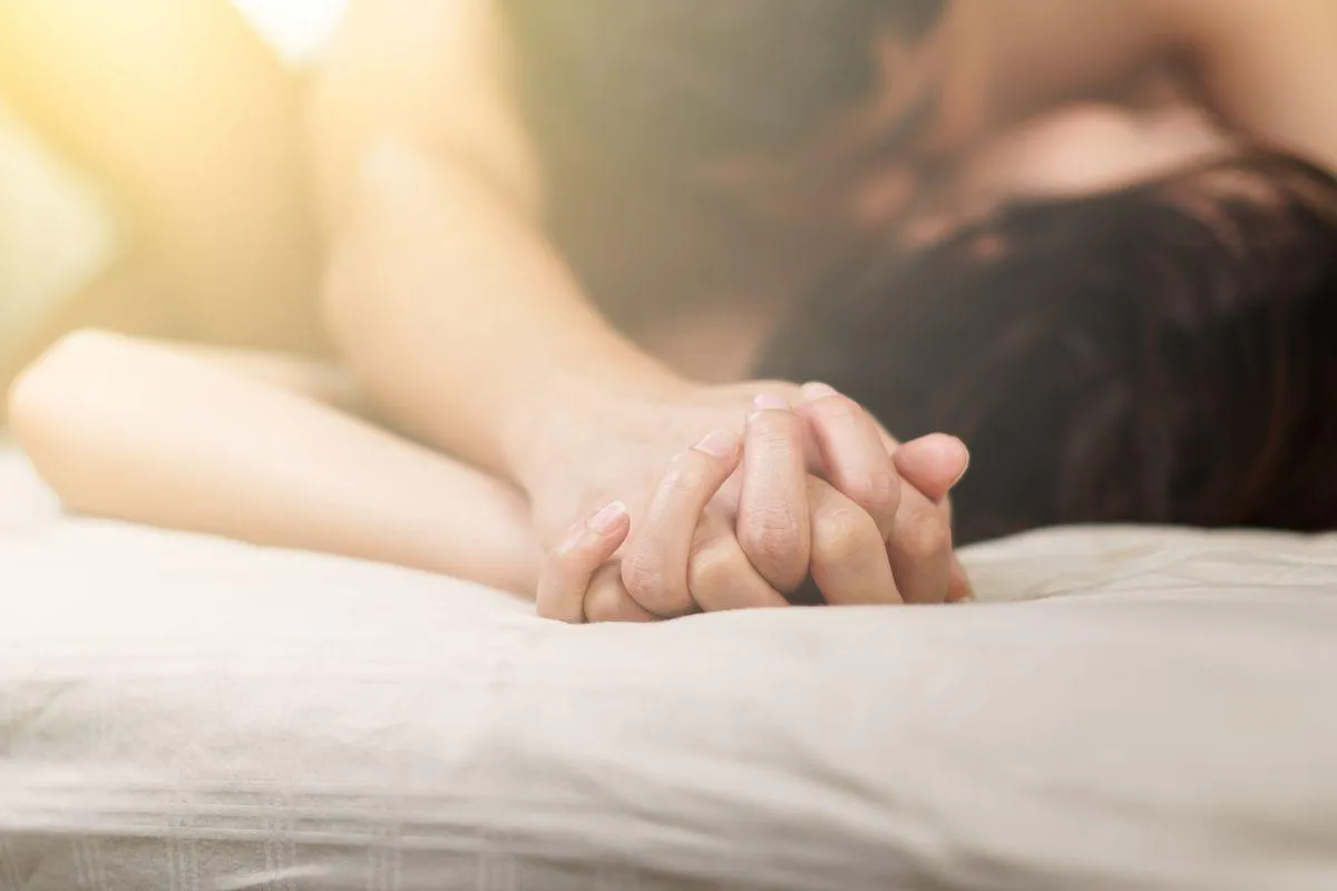 Two people of unclear sex and race holding hands, with one person laying on top of the other in bed.