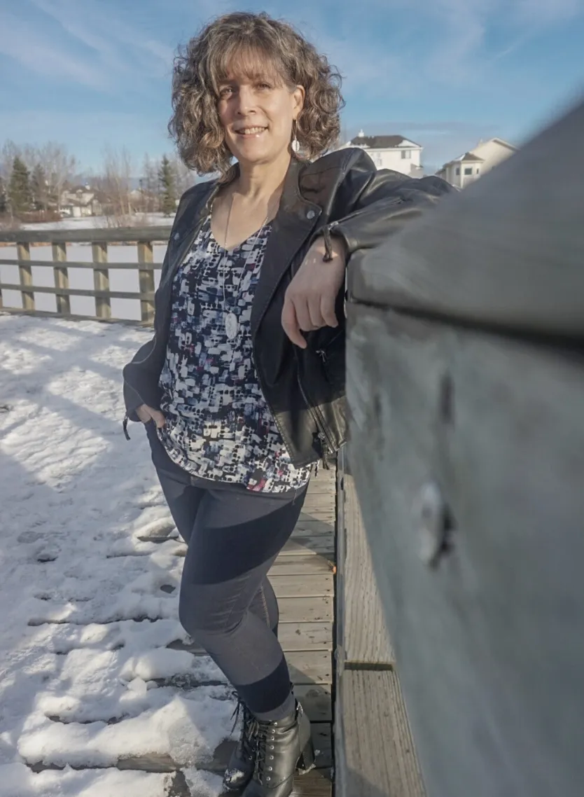 Leanne Chesser wearing blue jeans, a black leather jacket and a shirt with blue and maroon irregular shapes. She is standing with her left arm leaning on a wooden fence on a snow-covered pathway with houses in the background.