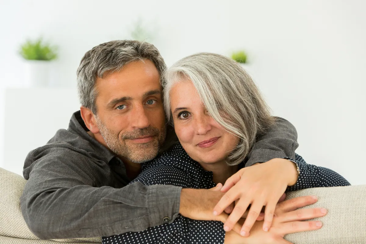 A middle-aged white woman with grey hair wearing a black, long-sleeved shirt with white polka dots and a middle-aged white man with grey hair wearing a grey, long-sleeved shirt, sitting next to each other and facing the camera while leaning against the back of a couch while on a date night.