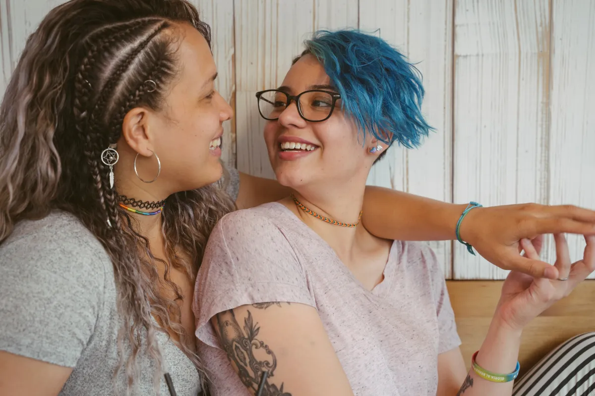 Two women of unclear race holding hands, sitting beside each other on a couch, looking at each other, smiling and talking.