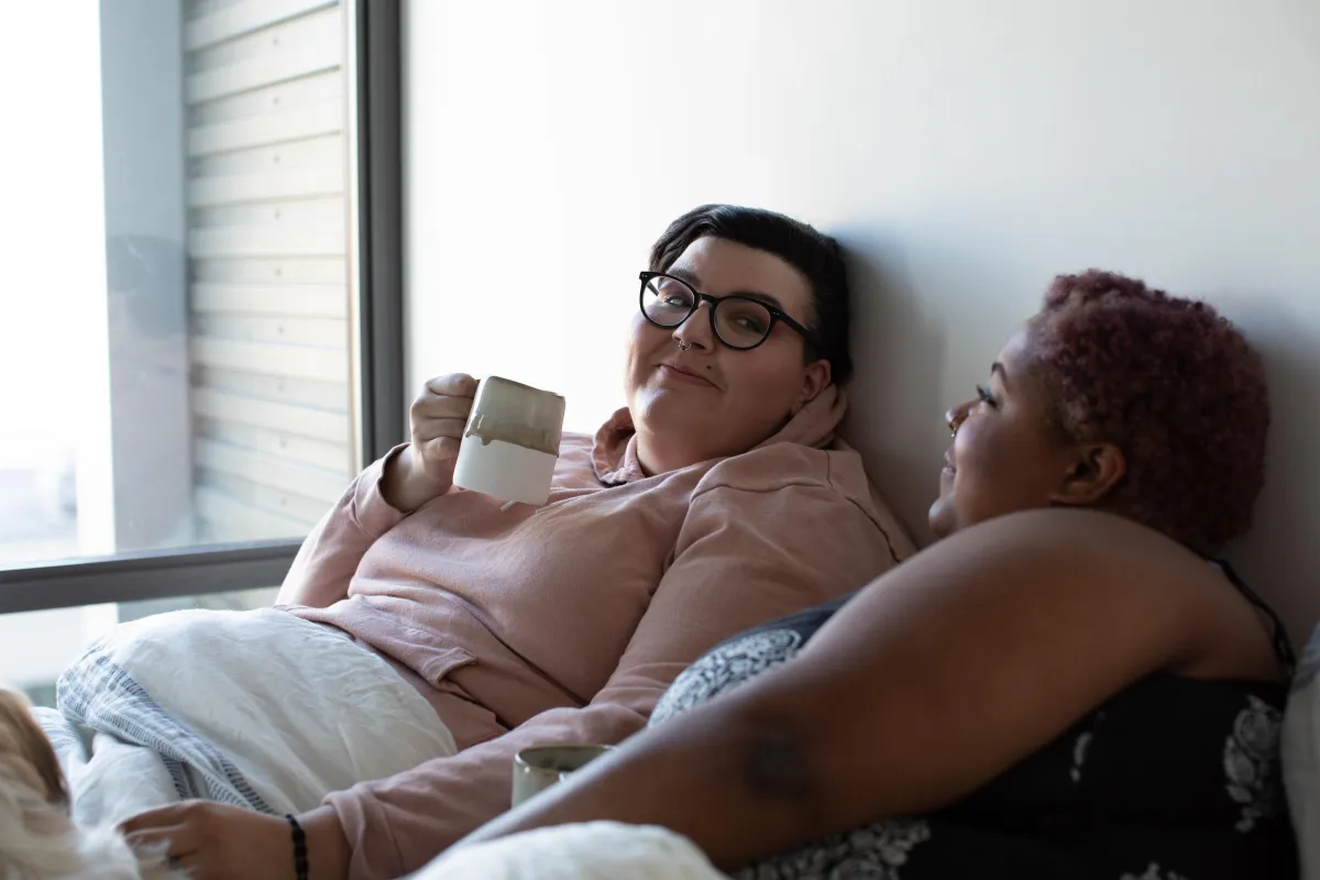 A lesbian couple sitting up side by side and under the covers in bed. One woman is white and is wearing a light pink shirt and black rimmed glasses and is holding a coffee cup. The other woman is Black and is laying on her side looking at her partner.