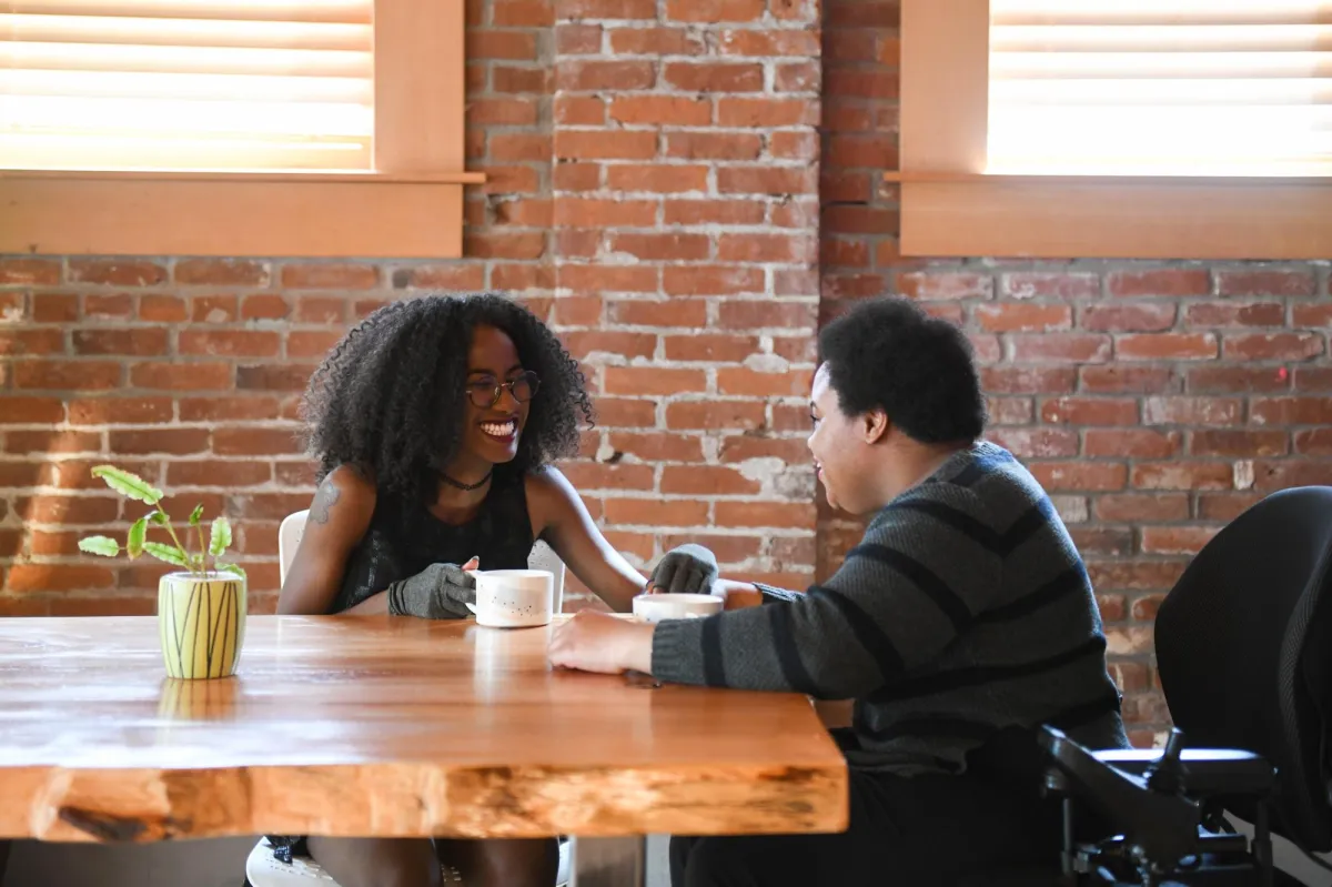 Two disabled Black people (a femme wearing compression gloves and a non-binary person in a power wheelchair that's partially in view) sit across from each other on a date night. Photo from Disabled And Here: https://affecttheverb.com/disabledandhere/
