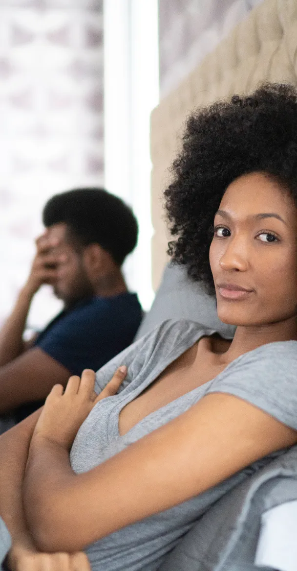 A disconnected couple on a date night. A black man wearing a navy blue shirt with his hand over his face sits far away from a black woman wearing a gray V-neck T-shirt with her arms crossed and looking away from the man toward the camera.