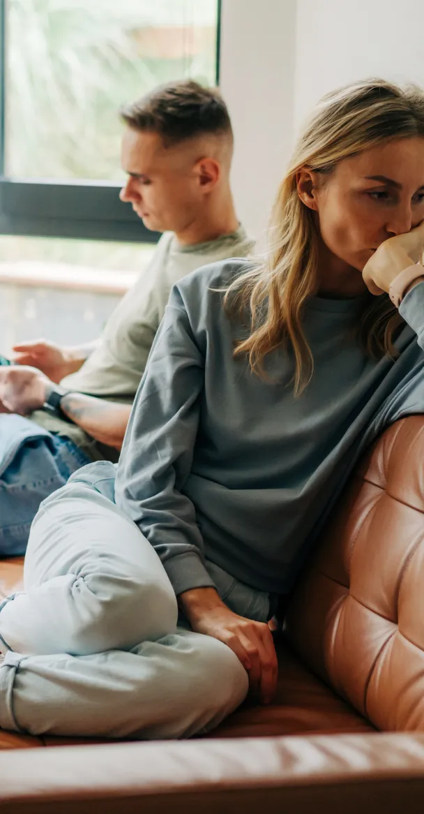 A white couple is disconnected on date night and sitting on a brown leather couch. The white man wearing a light sage green shirt is looking down at his phone while the white woman in a gray sweatshirt and light blue jeans sits turned away from him with her head leaning on her hand.