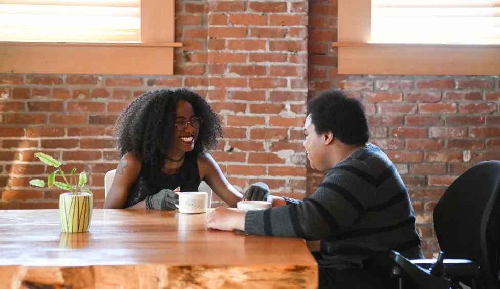 Two disabled Black people (a femme wearing compression gloves and a non-binary person in a power wheelchair that's partially in view) sit across from each other to create emotional connection. Photo from Disabled And Here: https://affecttheverb.com/disabledandhere/