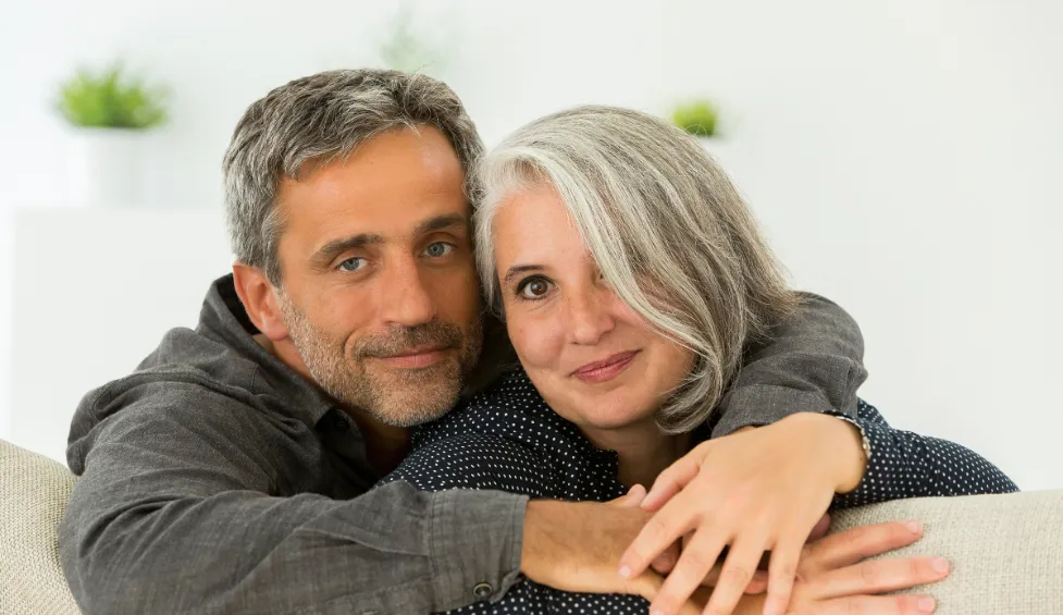 A middle-aged white woman with grey hair wearing a black, long-sleeved shirt with white polka dots and a middle-aged white man with grey hair wearing a grey, long-sleeved shirt, sitting next to each other and facing the camera while leaning against the back of a couch.