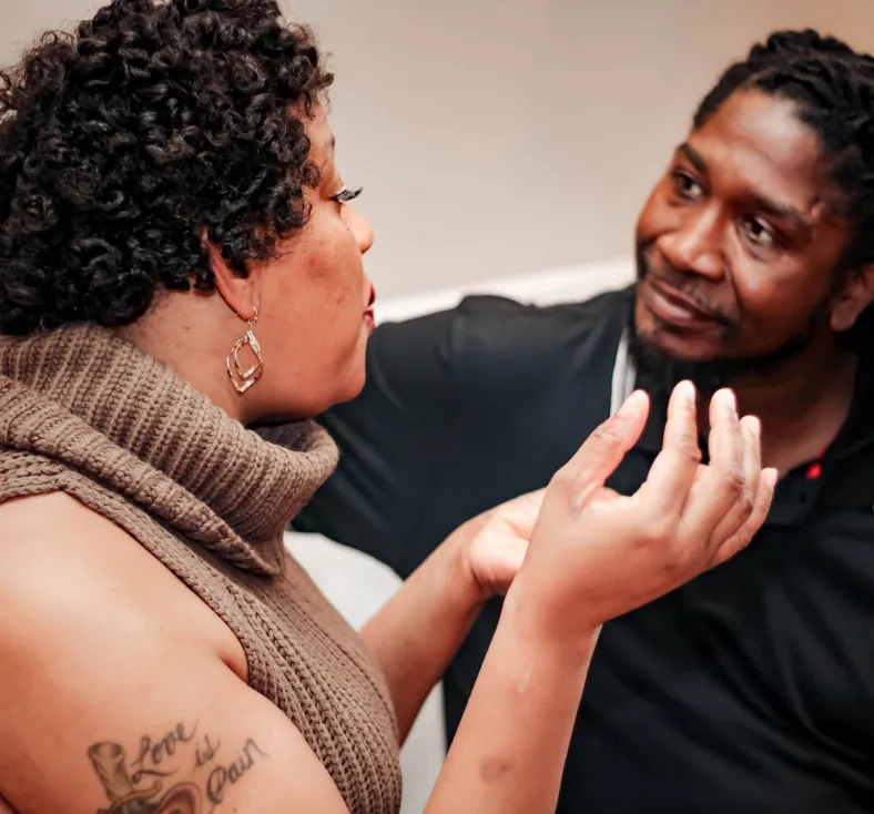 A black woman wearing a taupe sweater is talking to a black man wearing a black shirt. The man is looking at her intently, listening.