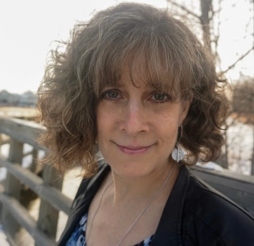 Head shot of Leanne Chesser standing in front of a wooden fence with trees and snow in the background and wearing a black leather jacket, a white shirt with blue geometric shapes and white and gray marble earrings and necklace.