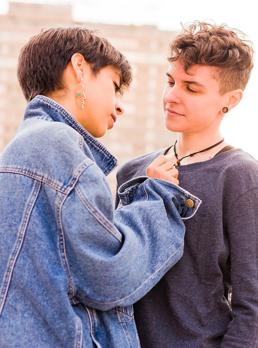 A non-binary couple standing outside facing each other, one wearing a blue jean jacket and the other partner wearing a dark gray shirt.