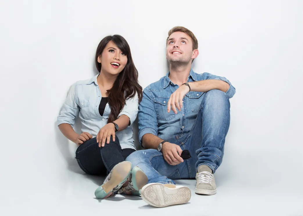 A connected couple (an Asian woman wearing a blue denim shirt and black jeans and a white man wearing a blue denim shirt and blue jeans) is sitting beside each other on the floor, shoulders and legs touching. They’re looking up and smiling, shifting intimacy issues and creating connection in their relationship.