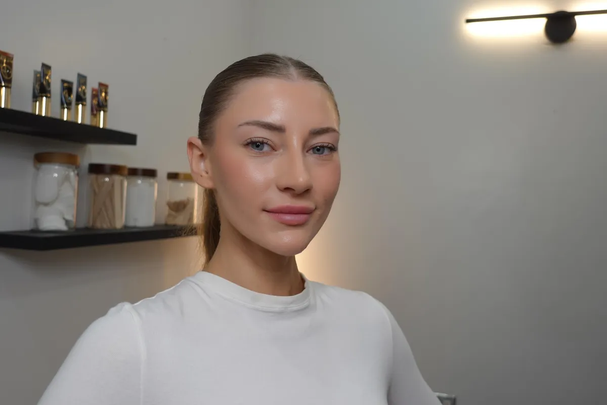 A woman holding makeup brushes standing next to a vanity mirror in a bright, stylish room.