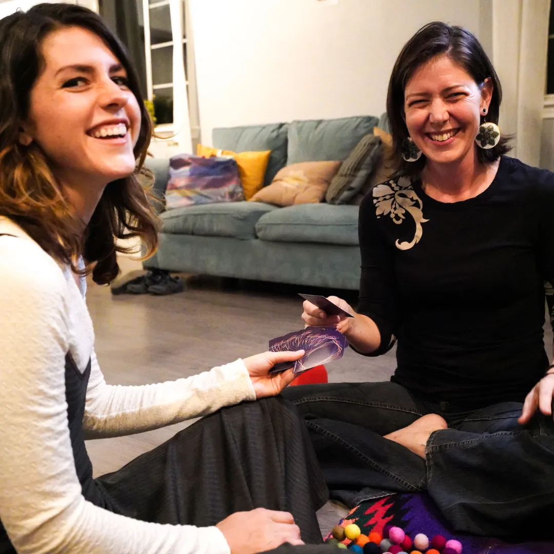 two women playing cards for connection in a comfortable living room. sitting on the floor, smiling and holding cards.