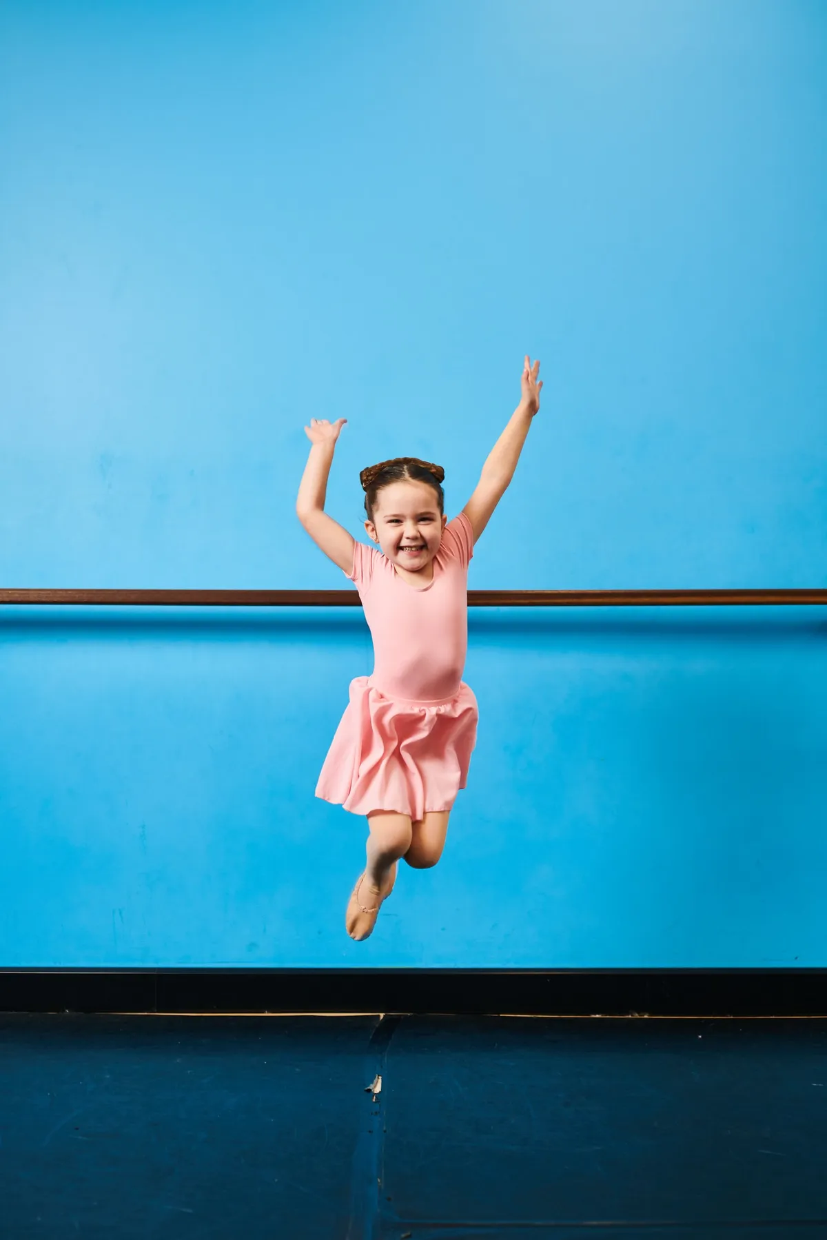 A little dancer in pink jumping high with her arms up, grinning