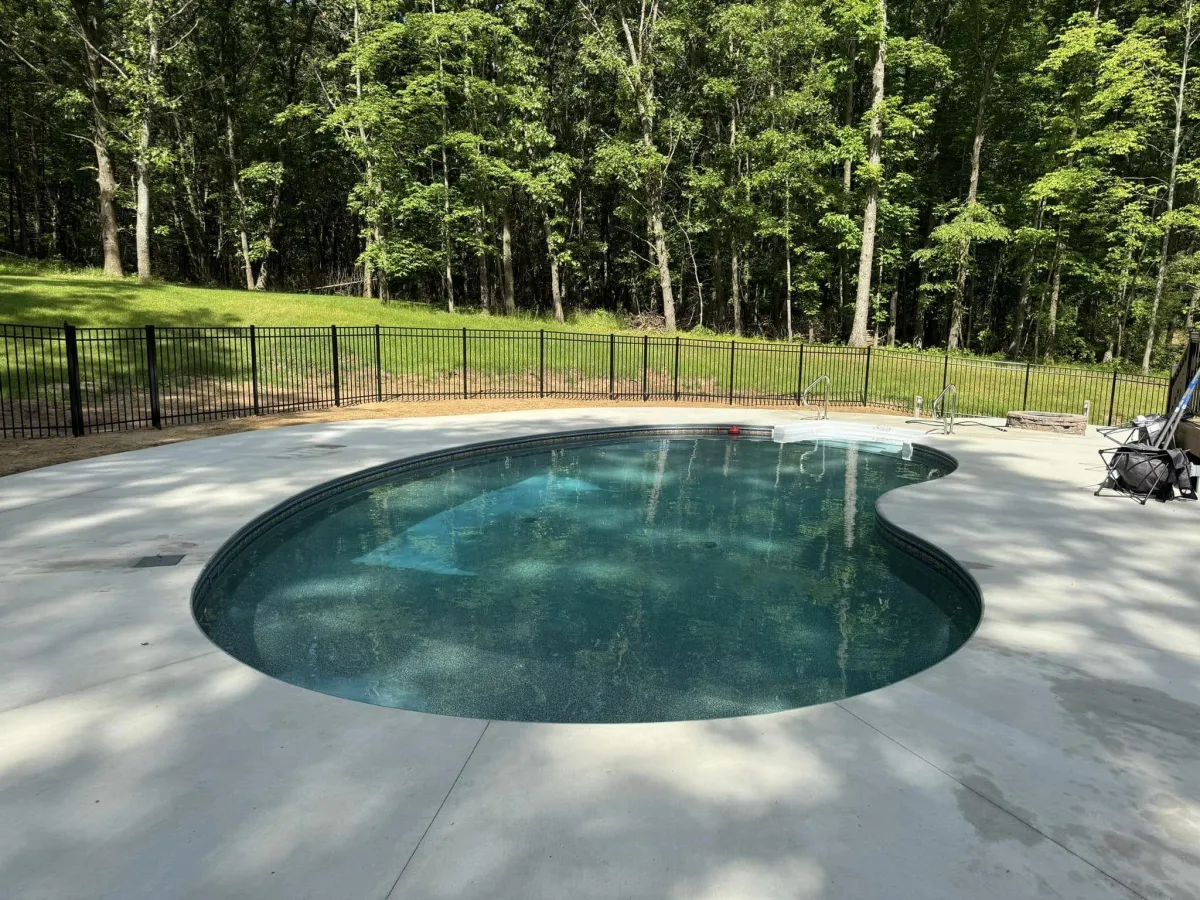 Finished custom inground pool filled with clear water, featuring a concrete pool deck and black metal fence surrounded by woods by Fizer Pools.