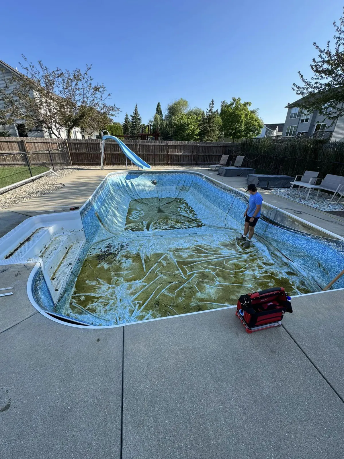 An empty inground swimming pool with a worn, stained vinyl liner in a residential backyard, before a liner replacement by Fizer Pools.