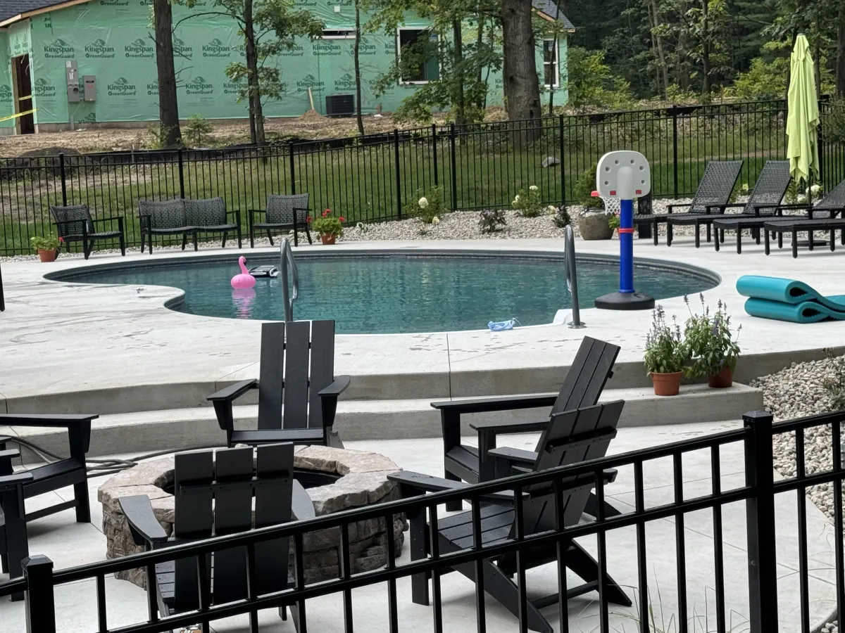 Completed crescent inground pool with concrete patio, fire pit, and black safety fence in Rockford, MI by Fizer Pools