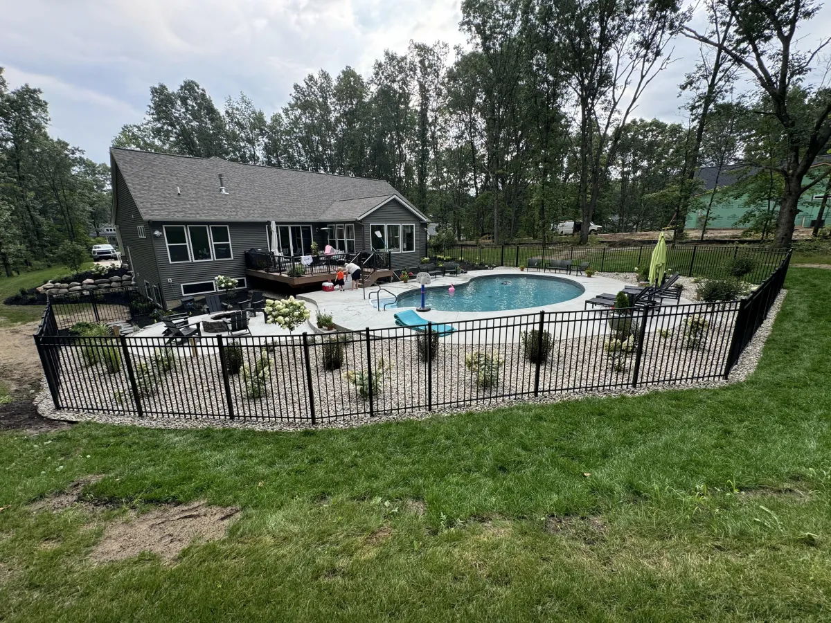 Wide backyard view of a completed custom crescent inground pool, concrete patio, landscaping, and safety fence in Rockford, MI by Fizer Pools