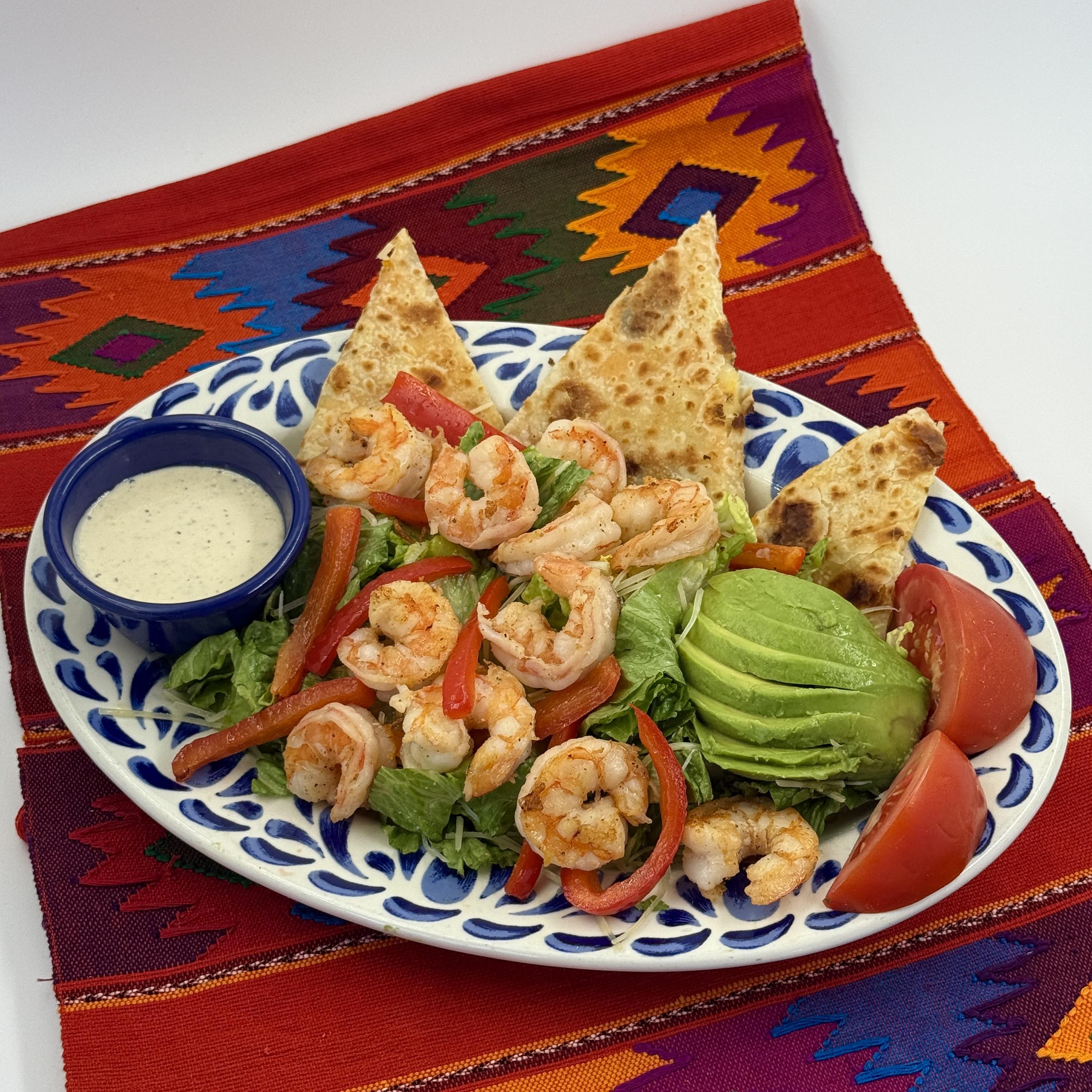 SHRIMP CAESAR SALAD Classic Caesar Salad originates just south of the border in Tijuana, Mexico. Crisp, refreshing romaine lettuce is the star of this beloved dish, providing a crunchy base that pairs perfectly with our rich, creamy Caesar dressing. Topped with seasoned grilled shrimp. This handcrafted dressing is a harmonious blend of garlic, tangy lemon juice, and Dijon mustard; all whisked together with fresh Parmesan cheese to create a perfect balance of flavors. A generous topping of crispy tortilla strips adds a satisfying crunch, while a final dusting of grated Parmesan cheese gives it a savory finish. Served with sliced avocado, tomato and quesadilla wedges.