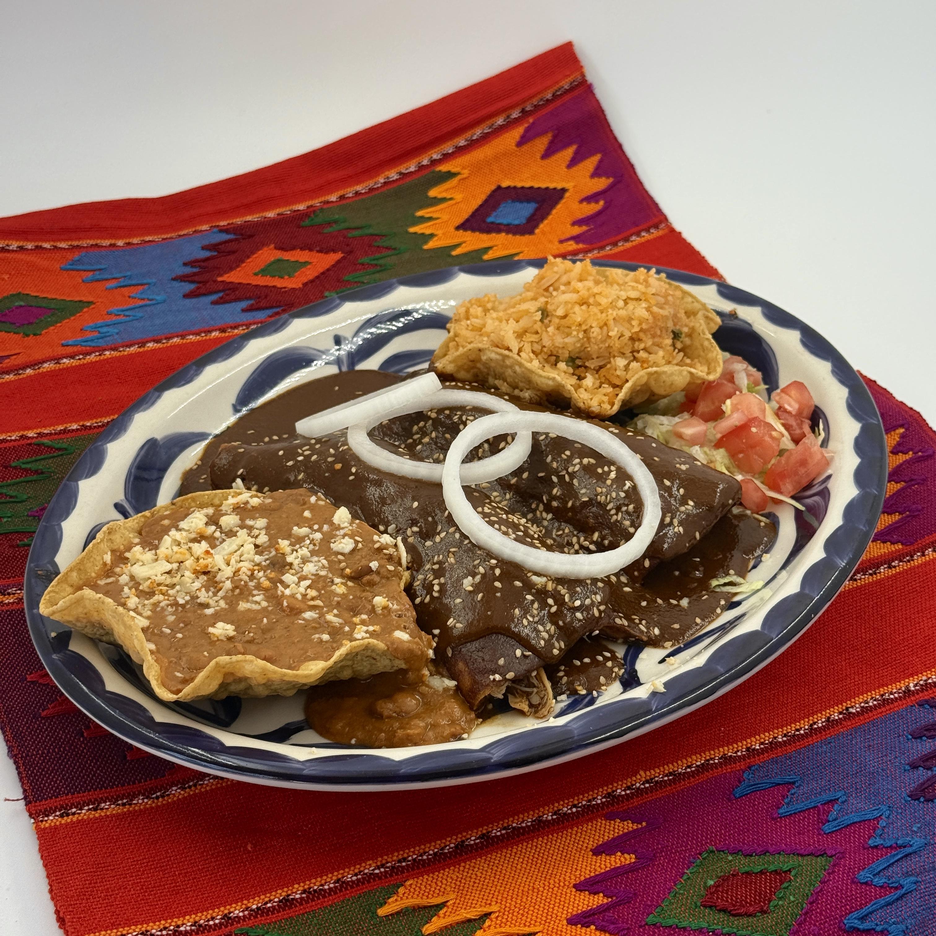 Chicken Mole Enchiladas, Pulled chicken enchiladas dressed in a homemade rich mole poblano sauce. Garnished with onions, crumbly cotija cheese, and a sprinkle of sesame seeds, this flavorful dish is accompanied by Mexican rice and Homemade beans for a satisfying meal.