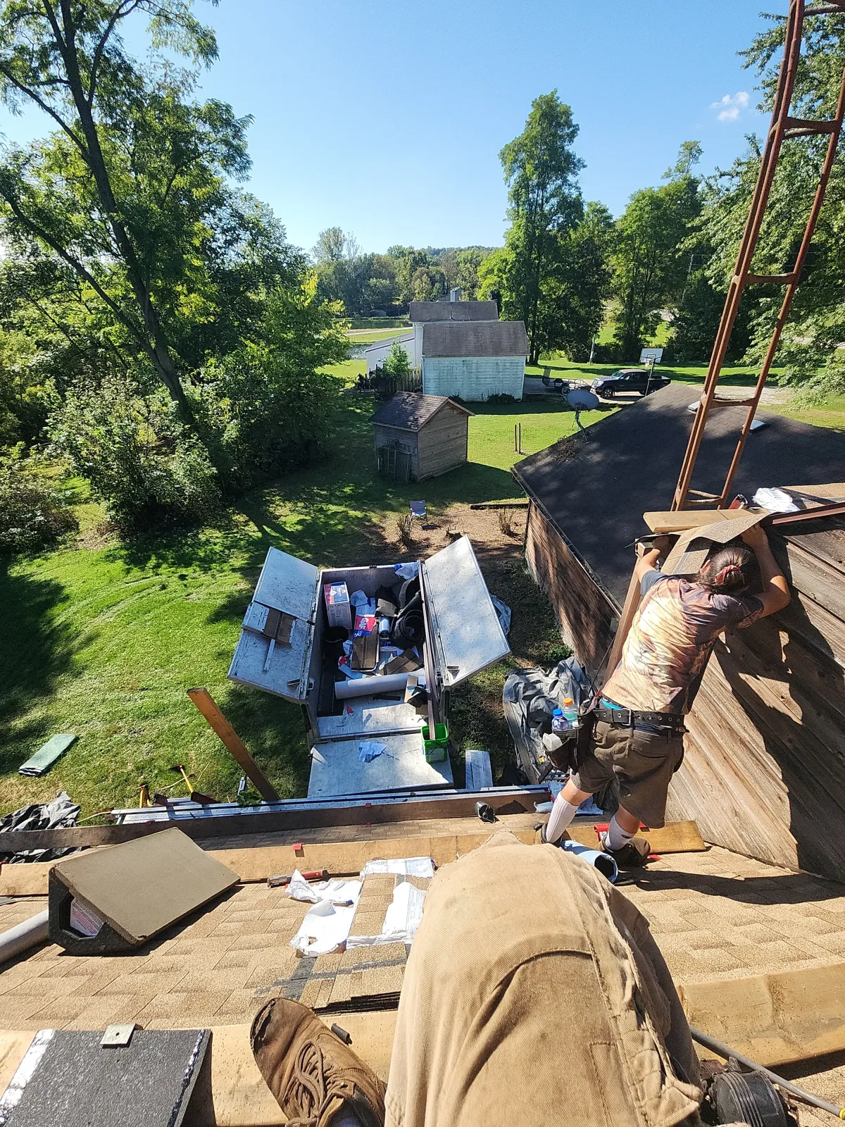 Weikles Roofing crew members actively working on a steep residential roof replacement project, with materials and equipment staged below on a beautiful sunny day. - Weikles Roofing