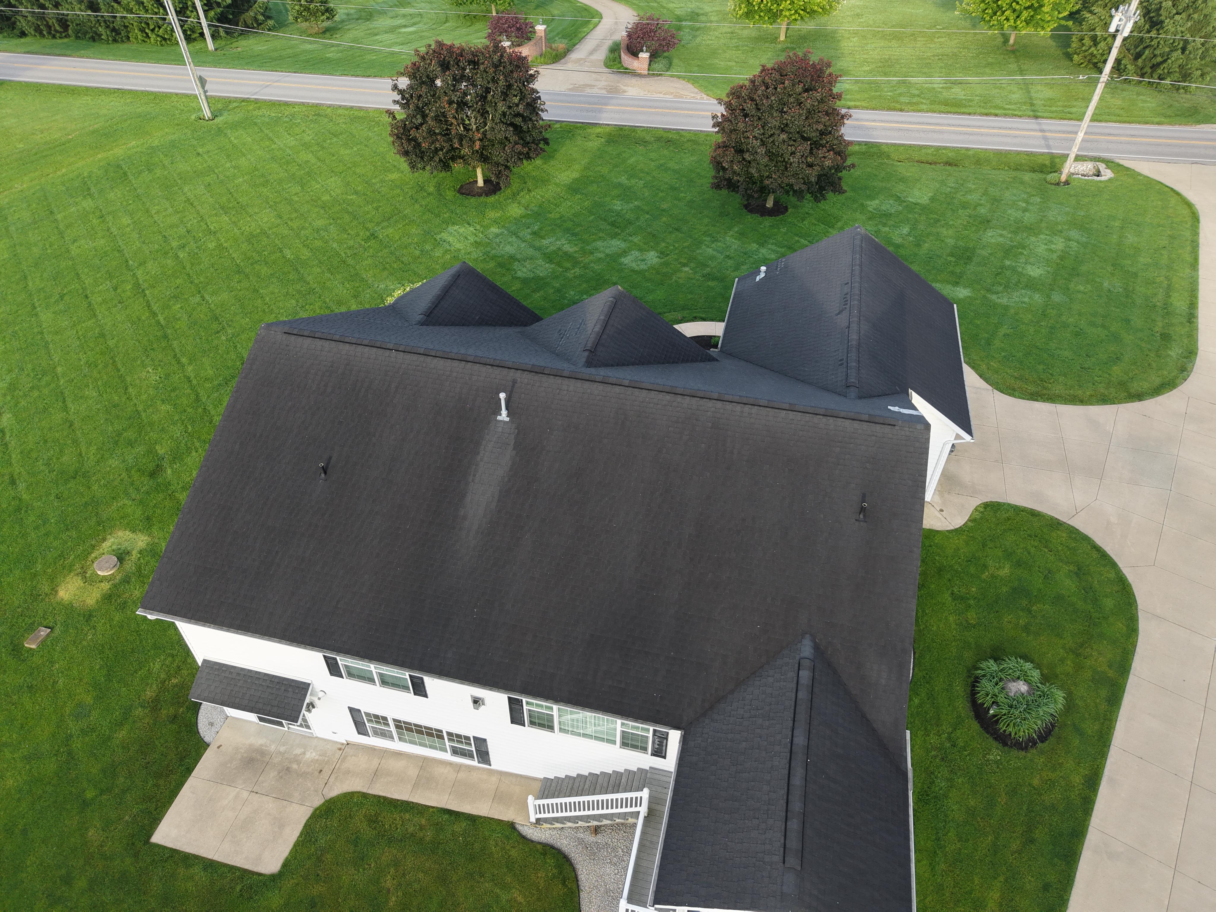 Aerial drone view of a large residential home featuring a recently installed dark charcoal asphalt shingle roof with multiple hip and gable sections, dormers, and clean ridge lines, set on a well-mani