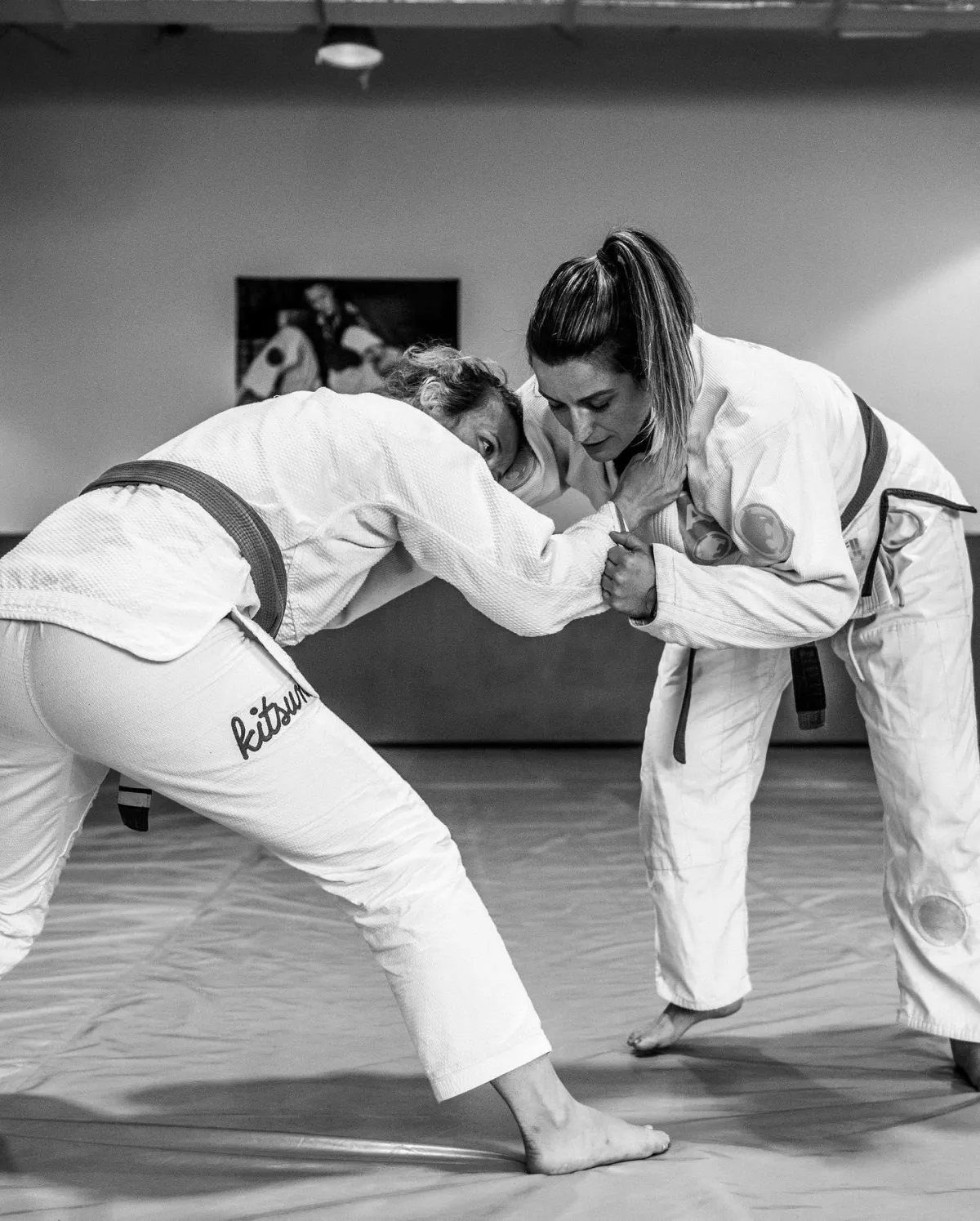 two women grappling with each other on the mat at the Renzo Gracie Academy of Jiu-Jitsu in Lake Worth. The women are wearing traditional jiu-jitsu uniforms (gi) and are fully engaged in their practice session. They are using a combination of grappling and groundwork techniques as they refine their jiu-jitsu skills. The gym has a spacious and well-lit training area, with mats covering the floor and motivational posters and banners on the walls. The atmosphere is one of discipline and concentration, as the women work to improve their jiu-jitsu proficiency. The image represents the inclusive and diverse community at the Renzo Gracie Academy, where women are encouraged and empowered to pursue their passion for martial arts.