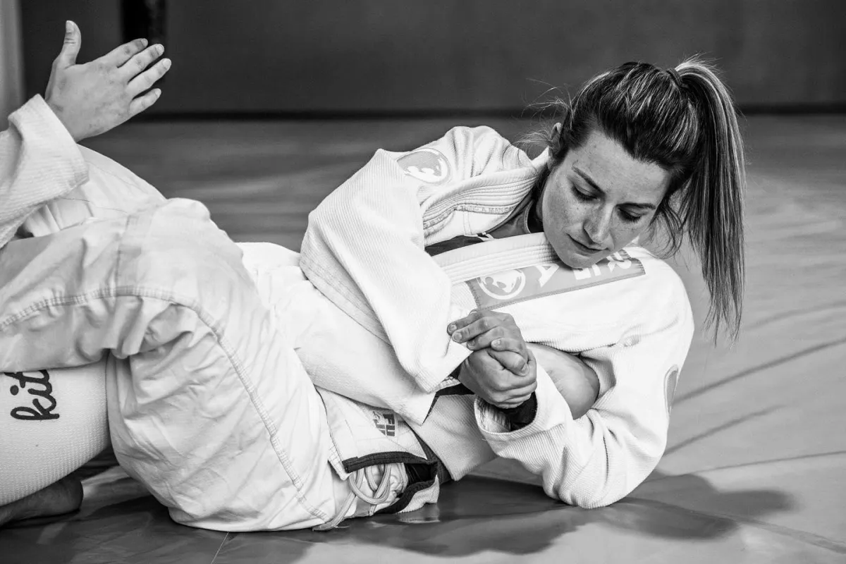 She is draped in a white Jiu-Jitsu gi and is surrounded by a group of similarly focused participants also training under the tutelage of the esteemed Renzo Gracie.  The Renzo Gracie Academy in Lake Worth is well-known in the world of Jiu-Jitsu and has produced world-class fighters. The image shows the dedication and hard work that goes into learning this artform as well as the commitment of the participants to better themselves physically and mentally. The mats of the Renzo Gracie Academy are pristine and the surroundings are clean and modern, evoking a sense of professionalism and discipline.