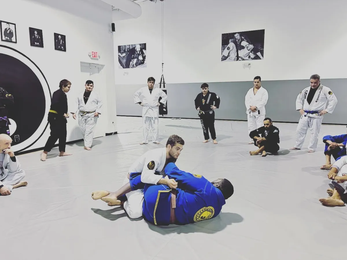 In the center of the image, two students are engaged in a jiu-jitsu practice session, rolling and grappling on a padded mat. The rest of the class is gathered around, observing the students' movements and techniques. The students are wearing traditional jiu-jitsu uniforms (gi), and are fully focused on their practice. The surrounding walls are adorned with motivational posters and the Renzo Gracie Jiu-Jitsu Academy logo. The atmosphere is one of intense concentration and mutual support, as the students work to improve their skills and techniques.
