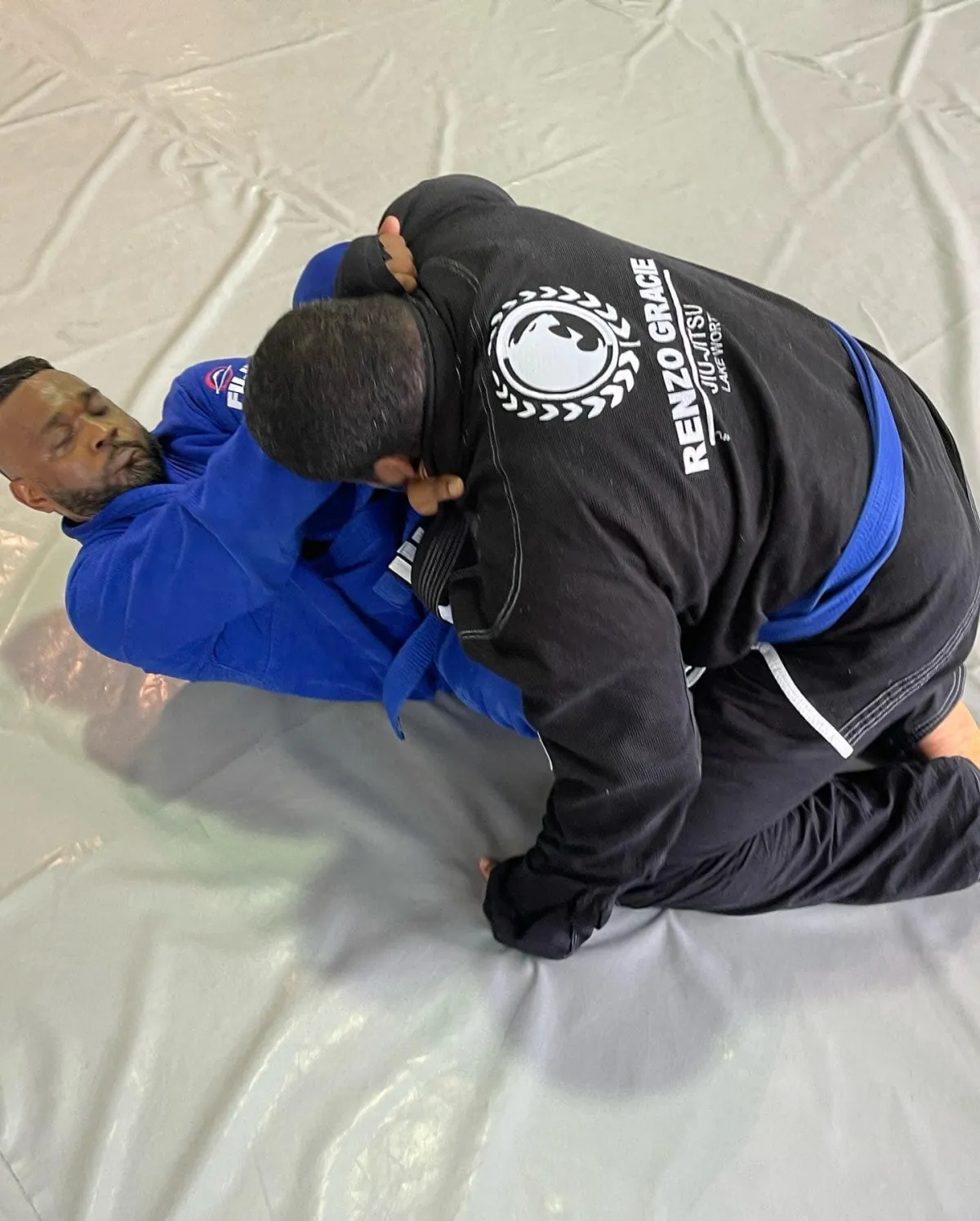 Shows two jiu-jitsu practitioners, both wearing blue belts, engaged in a practice session on the ground. They are wearing traditional jiu-jitsu uniforms (gi) and are fully focused on their movements and techniques. The two practitioners are using a combination of grappling and groundwork techniques, as they practice and refine their jiu-jitsu skills. The gym has a spacious and well-lit training area, with mats covering the floor and motivational posters and banners on the walls. The atmosphere is one of discipline and concentration, as the practitioners work to improve their jiu-jitsu proficiency.