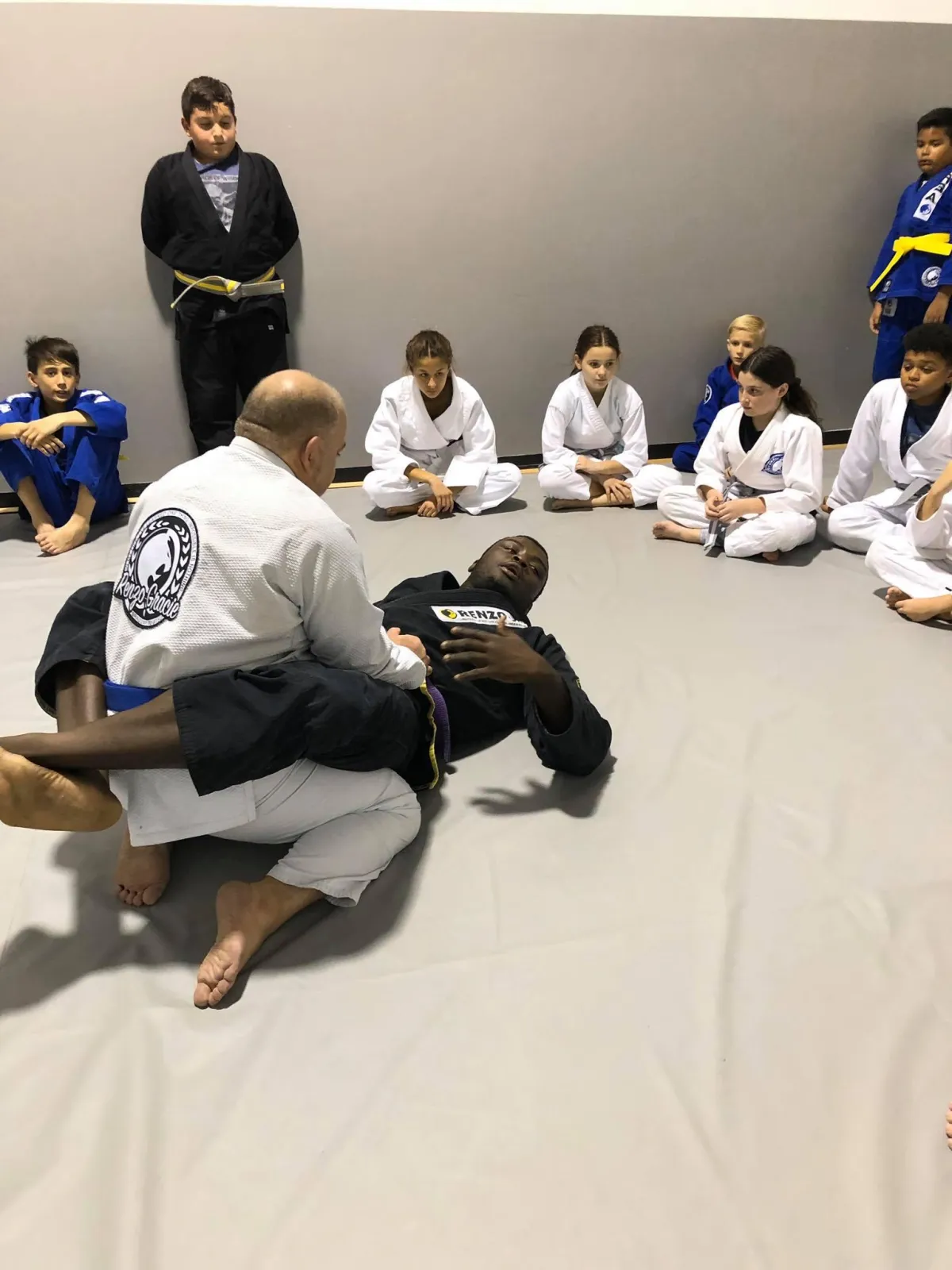 Two enthusiastic children attentively learning Brazilian Jiu-Jitsu techniques from Head Coach Adrian Alsina at Renzo Gracie Academy of Weston, known for empowering young minds through discipline and physical activity.