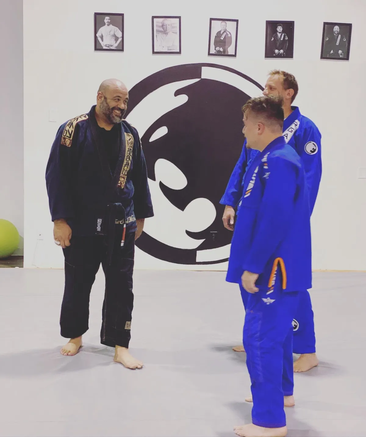 Head coach of the Renzo Gracie Jiu-Jitsu Academy of Lake Worth, Santos Caban, greeting two adult male students in the gym after a class. Caban is dressed in jiu-jitsu clothing and is shaking hands with the students, who are also wearing traditional jiu-jitsu uniforms (gi). The gym is spacious and brightly lit, with mats covering the floor and motivational posters and banners on the walls. The atmosphere is friendly and welcoming, as the coach and students exchange pleasantries and discuss their training session.