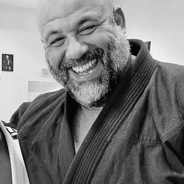 The image shows Santos Caban, the head professor at the Renzo Gracie Academy of Lake Worth, smiling for the camera. Caban is dressed in a traditional jiu-jitsu uniform (gi) and is standing in a spacious and brightly lit gym. The walls of the gym are adorned with motivational posters and banners featuring the Renzo Gracie Academy logo. Caban's smile reflects his warm and welcoming personality, which is reflected in the Academy's commitment to creating a supportive and inclusive environment for all students. The image represents the dedication and expertise of Caban as a jiu-jitsu instructor and the Renzo Gracie Academy's commitment to providing high-quality training to all of its students.