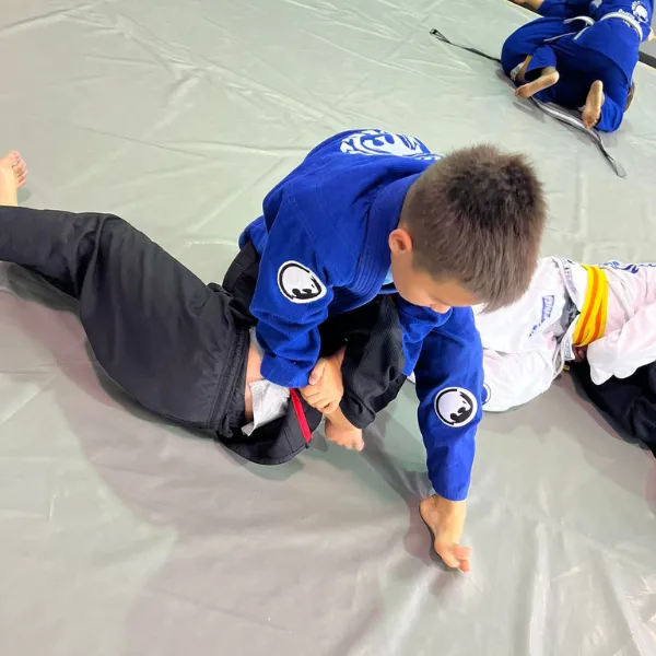 a young jiu-jitsu student practicing grappling on the mat in a class at the Renzo Gracie Academy of Lake Worth. The student is wearing a traditional jiu-jitsu uniform (gi) and is lying on the mat, fully engaged in the practice session. Other students can be seen in the background, also practicing their jiu-jitsu techniques. The gym has a spacious and well-lit training area, with mats covering the floor and motivational posters and banners on the walls. The atmosphere is one of discipline and focus, as the students work on developing their grappling skills under the guidance of their instructors. The image represents the commitment of the Renzo Gracie Academy to providing high-quality training to students of all ages and skill levels, and the dedication of the young student to mastering the techniques of jiu-jitsu alongside their peers.