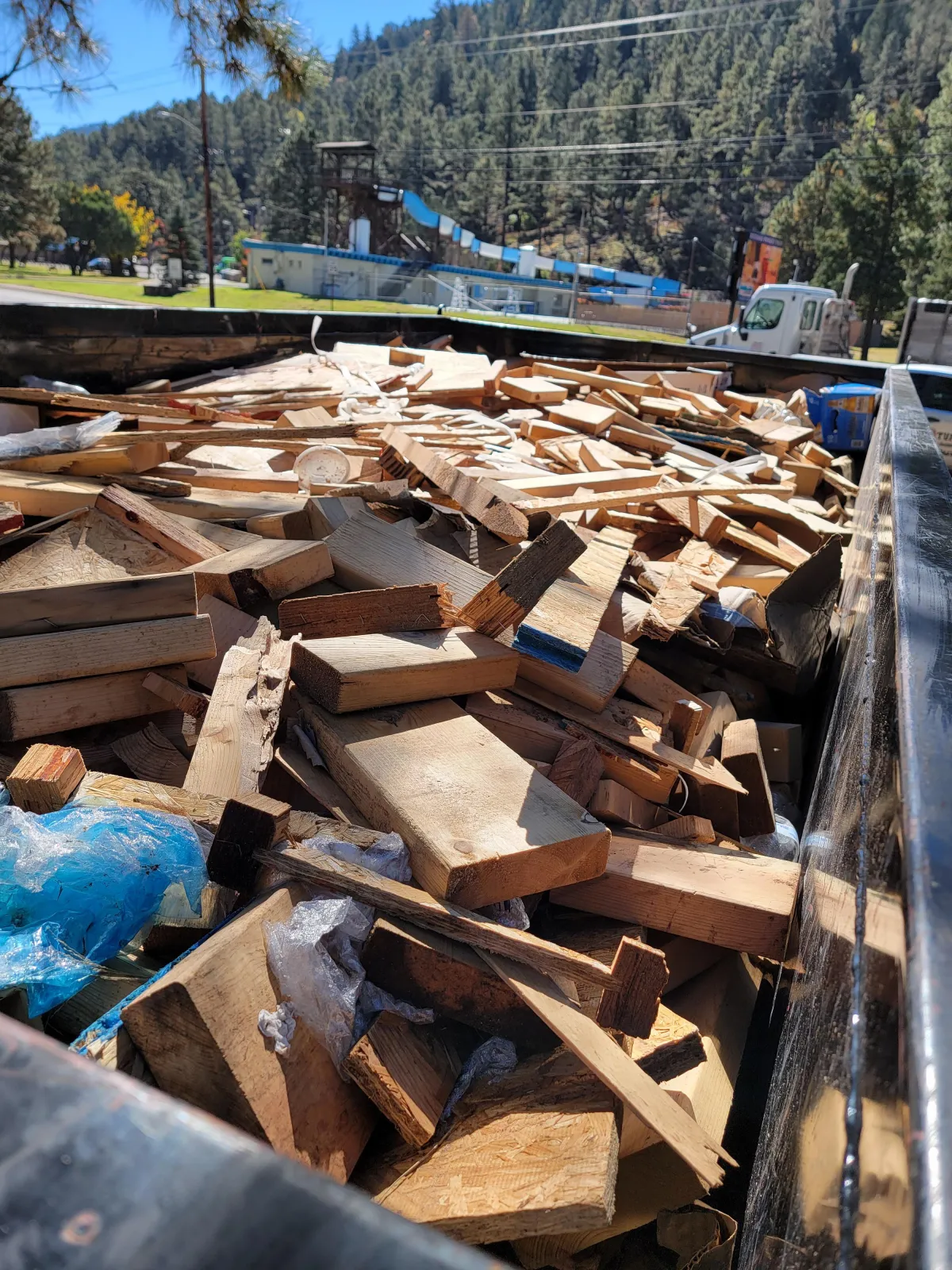 Open-top dumpster filled with clean wooden planks and lumber scraps near building and playground slide