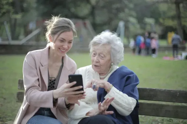 An elderly woman sits on a park bench and looks at a phone held by a younger woman during her companion care time provided by Sumner In-home Care and Preferred Care at Home