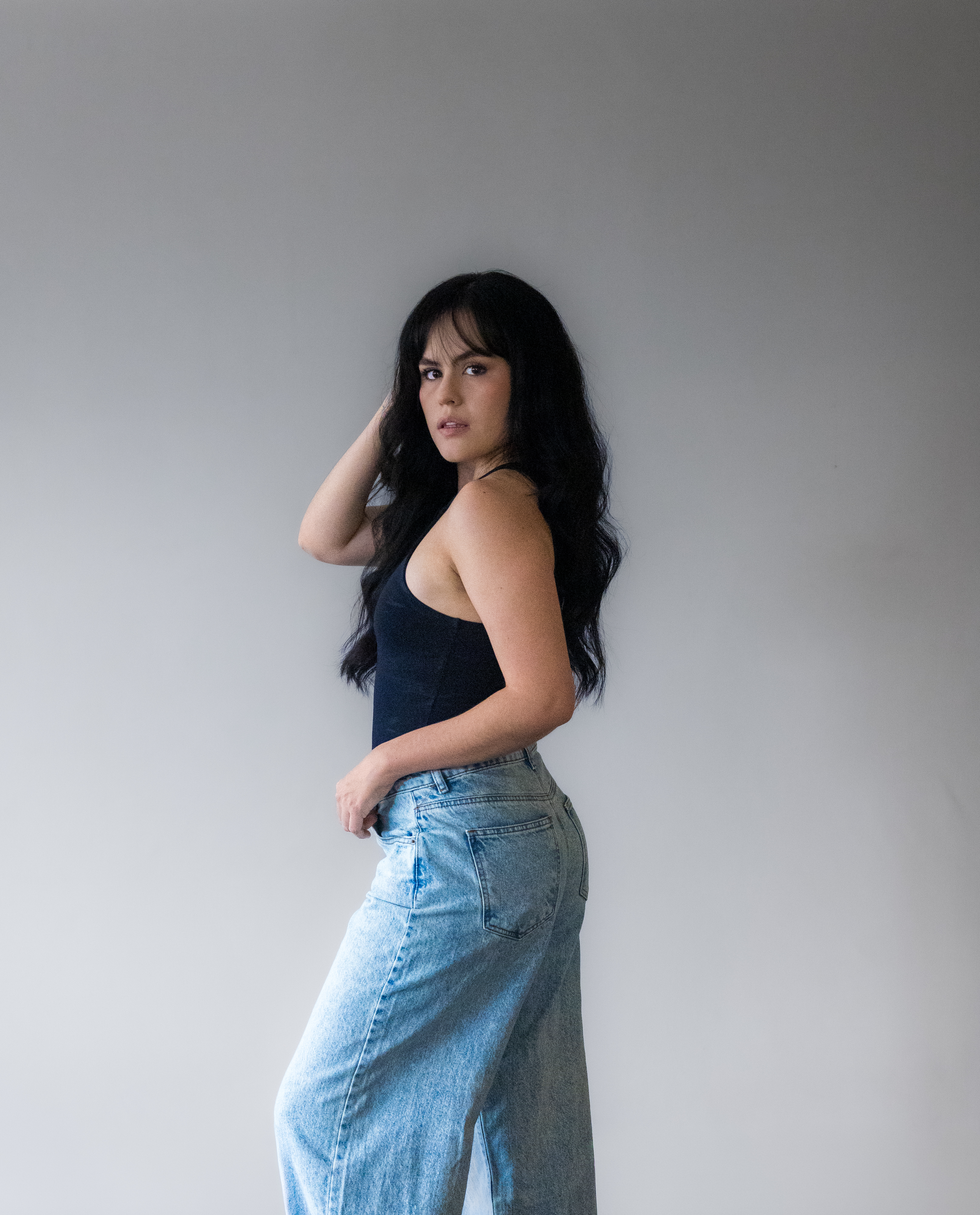 Woman posing sideways in casual jeans and a black top against a clean studio backdrop
