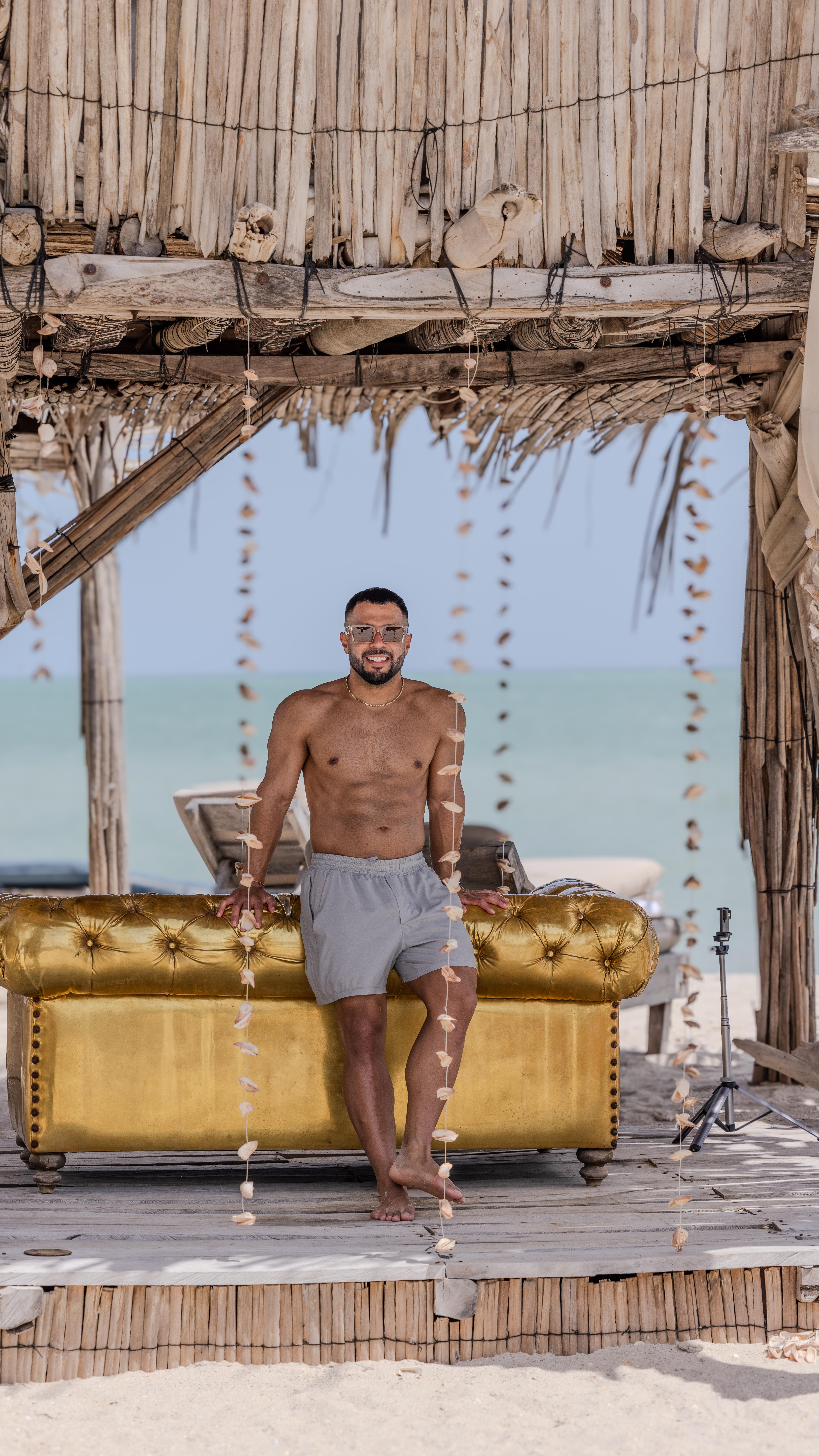 Man posing shirtless beside a rustic beach cabana with ocean view and wooden decor