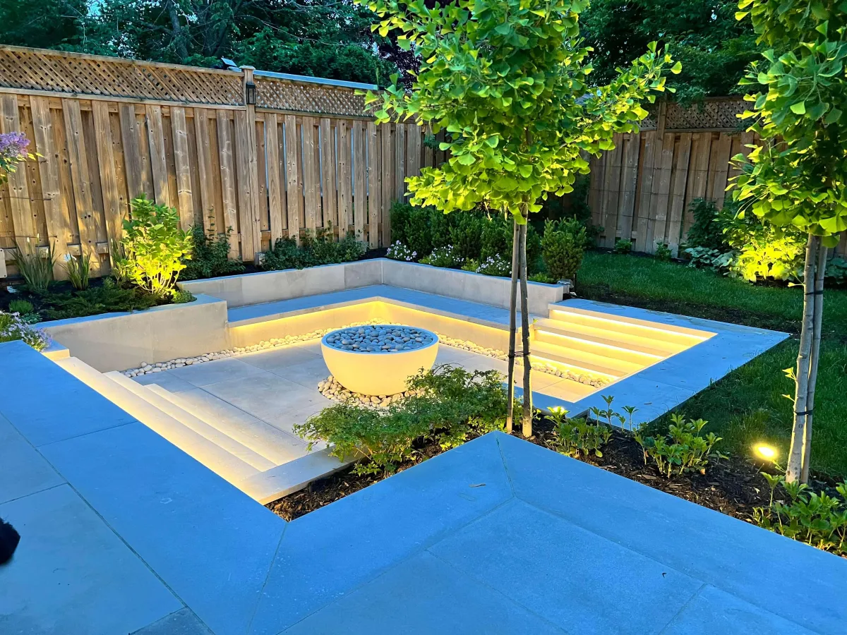 Modern garden design with a sunken seating area, ambient LED lighting, and a minimalist fire bowl centerpiece. Surrounded by lush greenery, stone paving, and wooden fencing, creating a stylish and functional outdoor oasis.