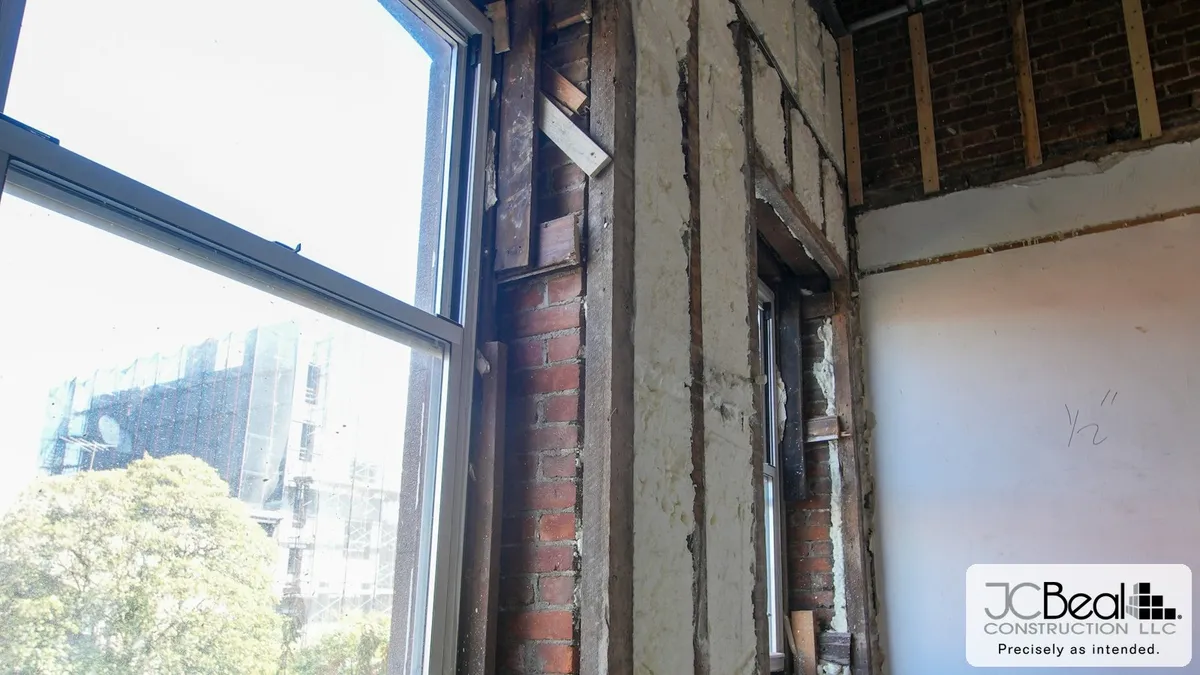 Opened wall cavity in a historic building showing original brick, exposed framing, and spray foam insulation next to a window overlooking the city