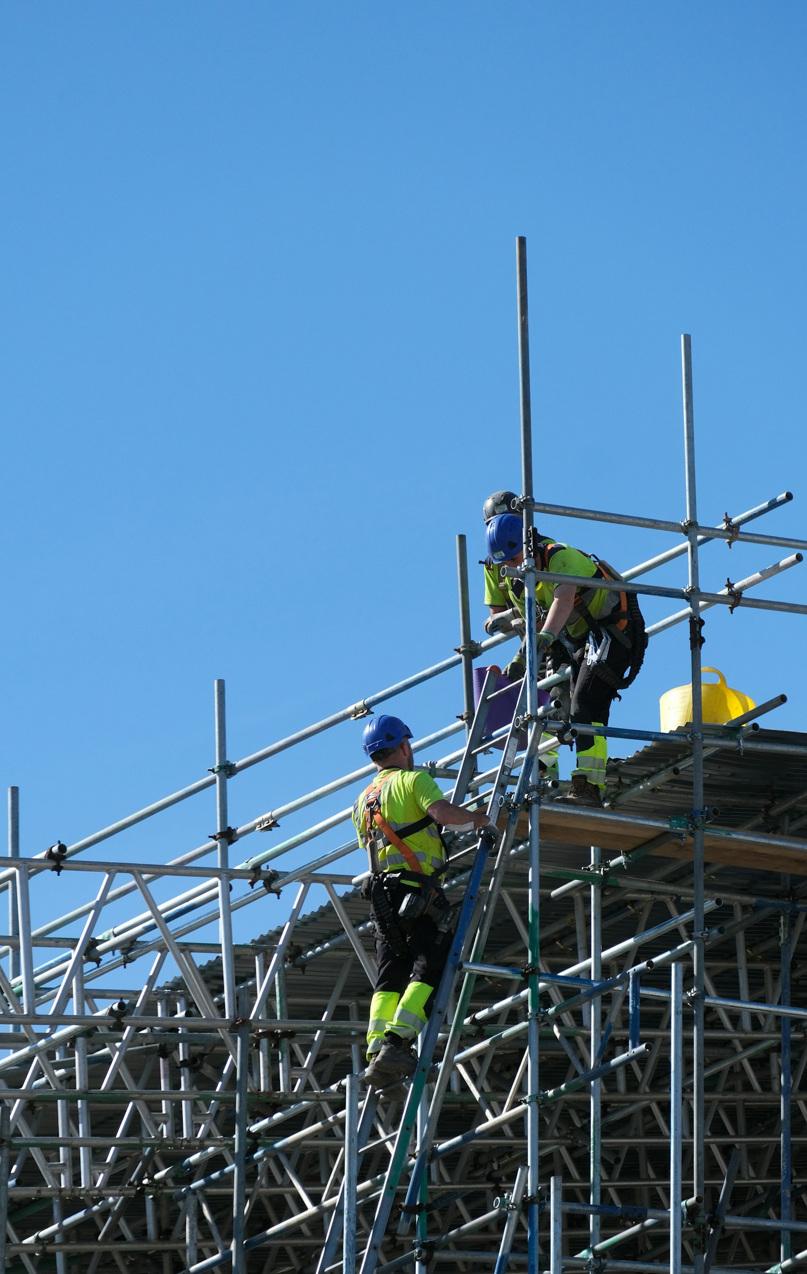 Two construction workers in safety harnesses and hard hats on scaffolding, stabilizing a building structure against a clear blue sky