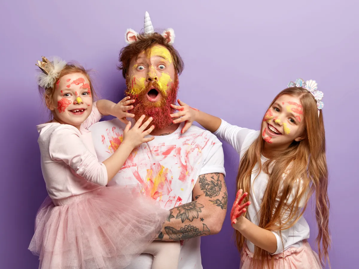 Father and two daughters laughing and playing together, covered in colourful paint, expressing playful family connection